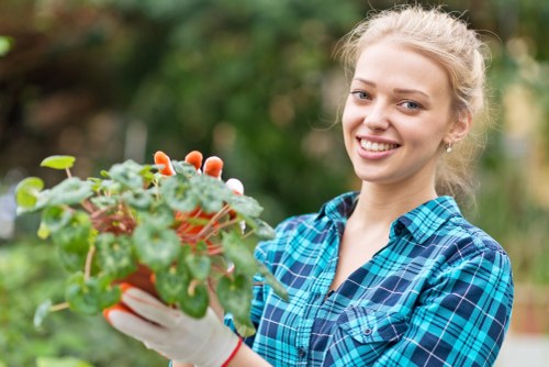 A young woman with blonde hair tied back, smiling and wearing a blue plaid shirt and white gardening gloves, holds a potted plant with green and reddish-pink foliage in an outdoor garden setting. The background features a blurred natural landscape with green trees and shrubbery, suggesting a well-maintained garden or backyard space. The scene is illuminated by natural daylight, indicating fair weather conditions, and reflects outdoor gardening activities typical in residential gardens in Gunnersbury, London. The image highlights the care and attention involved in garden maintenance supported by professional gardening services in the local area, with a focus on healthy plant care, soil management, and garden aesthetics.